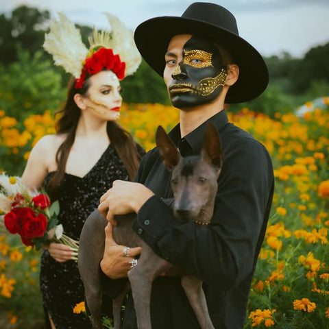 Man with face paint holding a cute dog, calm during Halloween in a field of marigolds, with a woman in costume holding flowers
